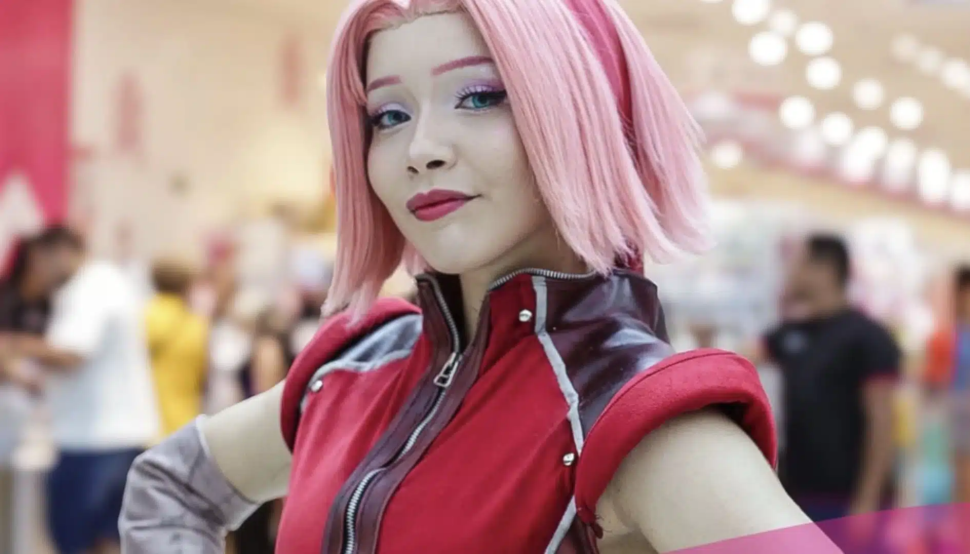 Close de uma cosplayer caracterizada como a personagem Sakura Haruno, de Naruto. Ela possui cabelos curtos cor-de-rosa, olhos claros e veste o traje clássico vermelho com detalhes em cinza. A jovem sorri levemente para a câmera em um ambiente de evento com fundo desfocado.