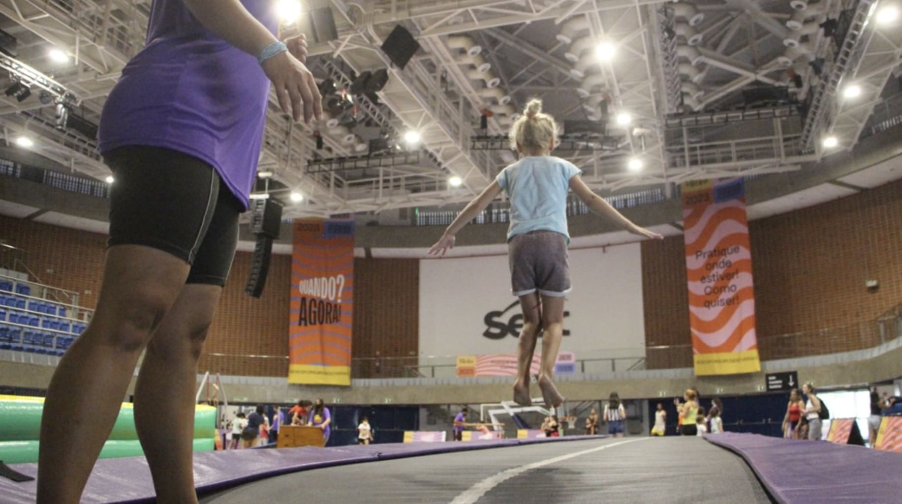 Criança de costas saltando em uma pista acrobática inflável dentro de um ginásio do Sesc. Uma instrutora de camiseta roxa observa em primeiro plano, à esquerda.