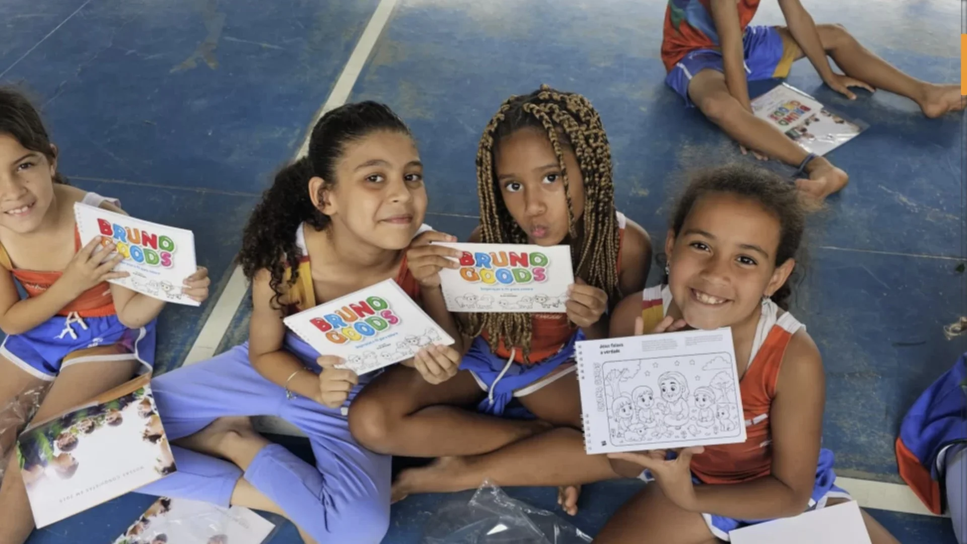 Quatro crianças sentadas no chão de uma quadra azul, sorrindo e segurando cadernos de colorir da marca "Bruno Goods". Uma das crianças mostra uma página interna com um desenho religioso para pintar. Elas vestem uniformes escolares laranjas e azuis.