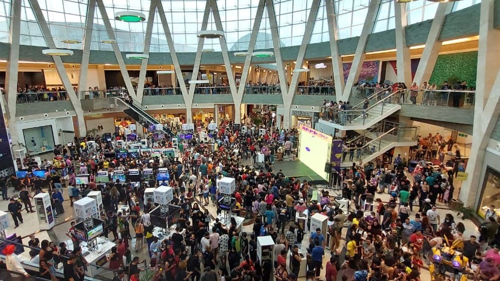 Vista aérea de um grande saguão de shopping lotado de pessoas durante uma exposição de videogames, com dezenas de estações de jogos, telões e uma arquitetura moderna com teto de vidro.