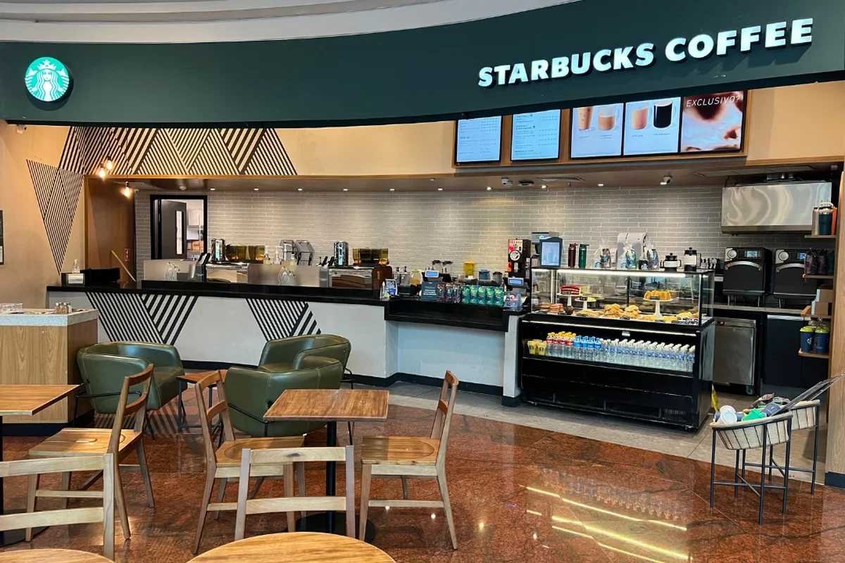 A loja possui um balcão moderno com painéis em padrões geométricos a preto e branco. Por trás do balcão, vê-se a área de preparação com máquinas de café e o menu digital fixado na parte superior.