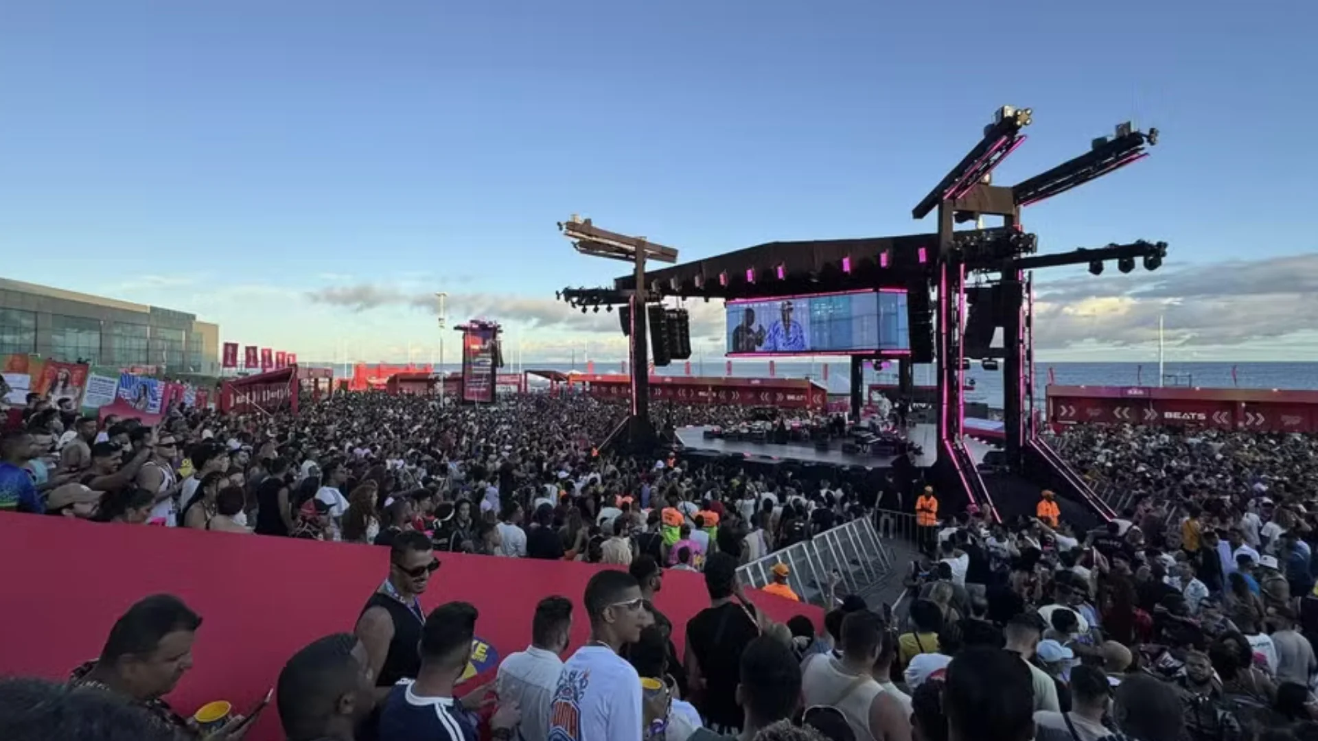 Vista panorâmica de uma grande multidão reunida em um evento ao ar livre durante o dia, em frente a um palco moderno com telões de LED. O local parece ser à beira-mar sob um céu claro.