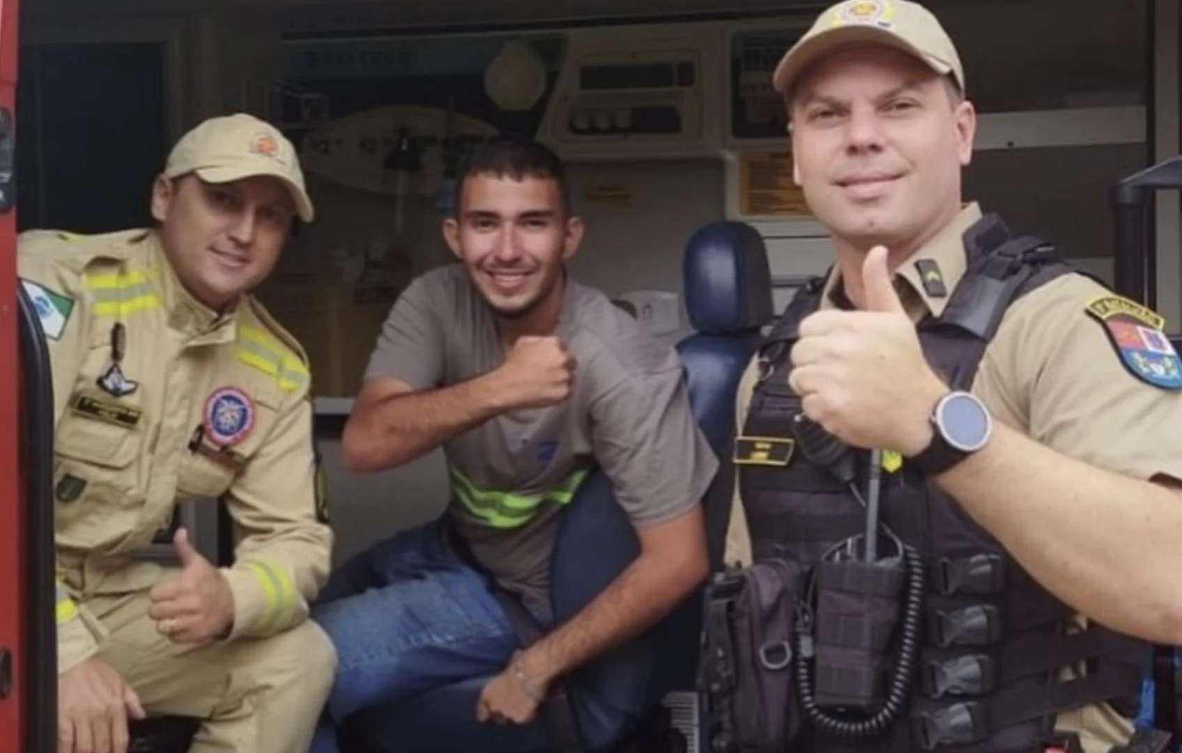 Três homens sorrindo dentro de uma ambulância: um jovem sentado ao centro ladeado por dois socorristas uniformizados que fazem sinal de positivo.