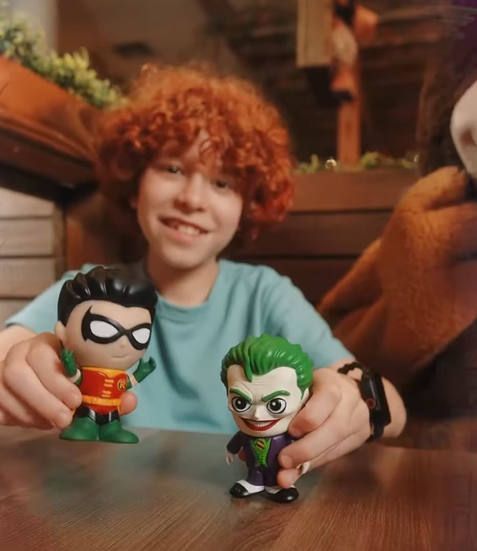 Uma criança sorridente com cabelos cacheados ruivos segura dois bonecos colecionáveis sobre uma mesa: o herói Robin na mão direita e o vilão Coringa na mão esquerda. Os bonecos têm o mesmo estilo 'chibi' da imagem anterior, com cabeças grandes e traços caricatos.