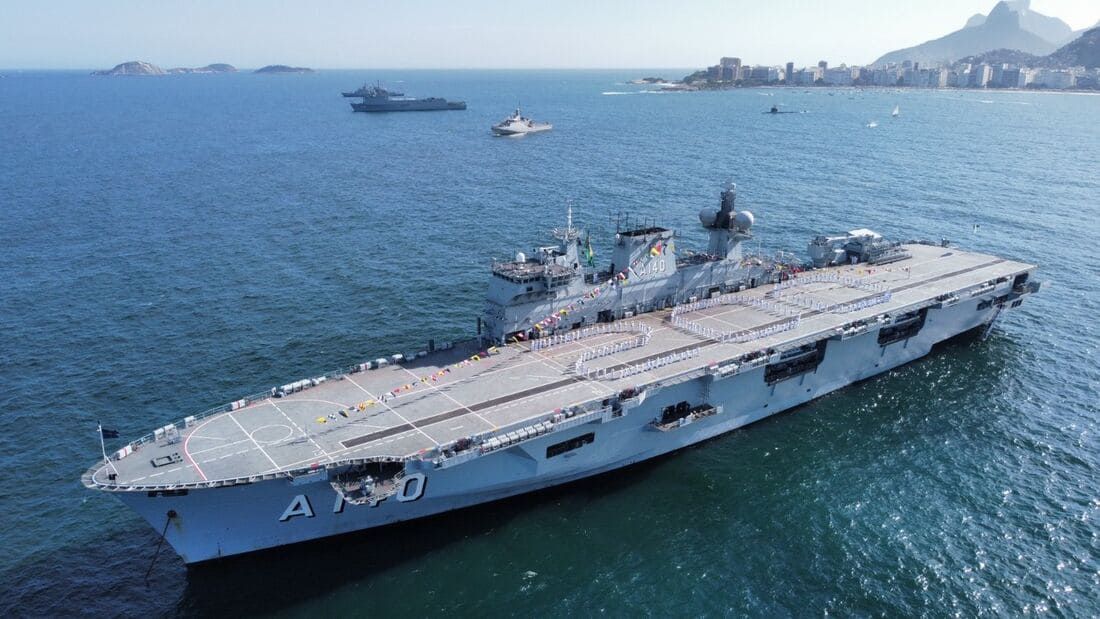 Vista aérea do NAM Atlântico (A140), o maior navio da Marinha do Brasil, navegando em águas azuis com a orla do Rio de Janeiro ao fundo. O convés de voo está ocupado por tripulantes em formação, e outras embarcações menores são visíveis no horizonte.