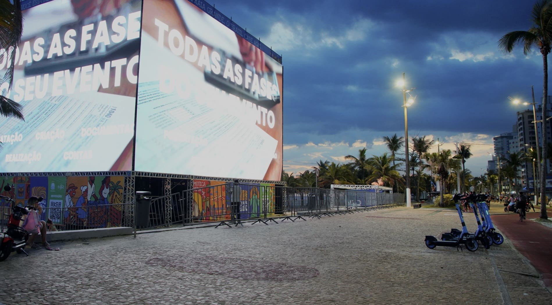 Vista ampla da orla de Praia Grande ao entardecer, destacando um grande cubo de LED iluminado, o calçadão de pedras e patinetes elétricos estacionados sob um céu nublado.