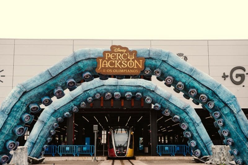 Fotografia frontal da entrada de um túnel de transporte público. Sobre a entrada, há uma escultura gigante de dois tentáculos azuis com ventosas que formam um arco. No topo do arco, está fixada a placa de madeira com o logotipo 'Disney Percy Jackson e os Olimpianos'. Um bonde moderno está parado no centro do trilho, saindo do túnel.