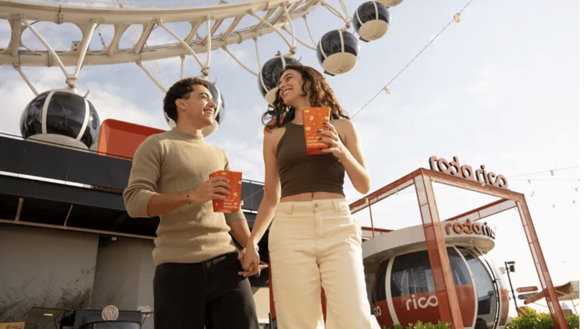 Um casal jovem caminha de mãos dadas sorrindo, segurando baldes de pipoca laranjas, em frente à estrutura da Roda Rico. Ao fundo, veem-se as cabines brancas e o logotipo da atração.