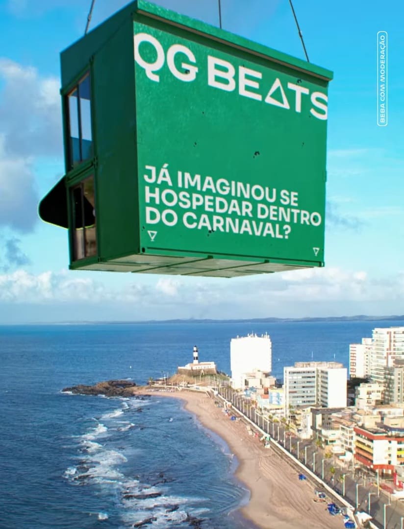 Vista aérea da orla de Salvador com o Farol da Barra ao fundo. Em primeiro plano, um contêiner verde suspenso no ar com o texto "QG BEATS: JÁ IMAGINOU SE HOSPEDAR DENTRO DO CARNAVAL?".