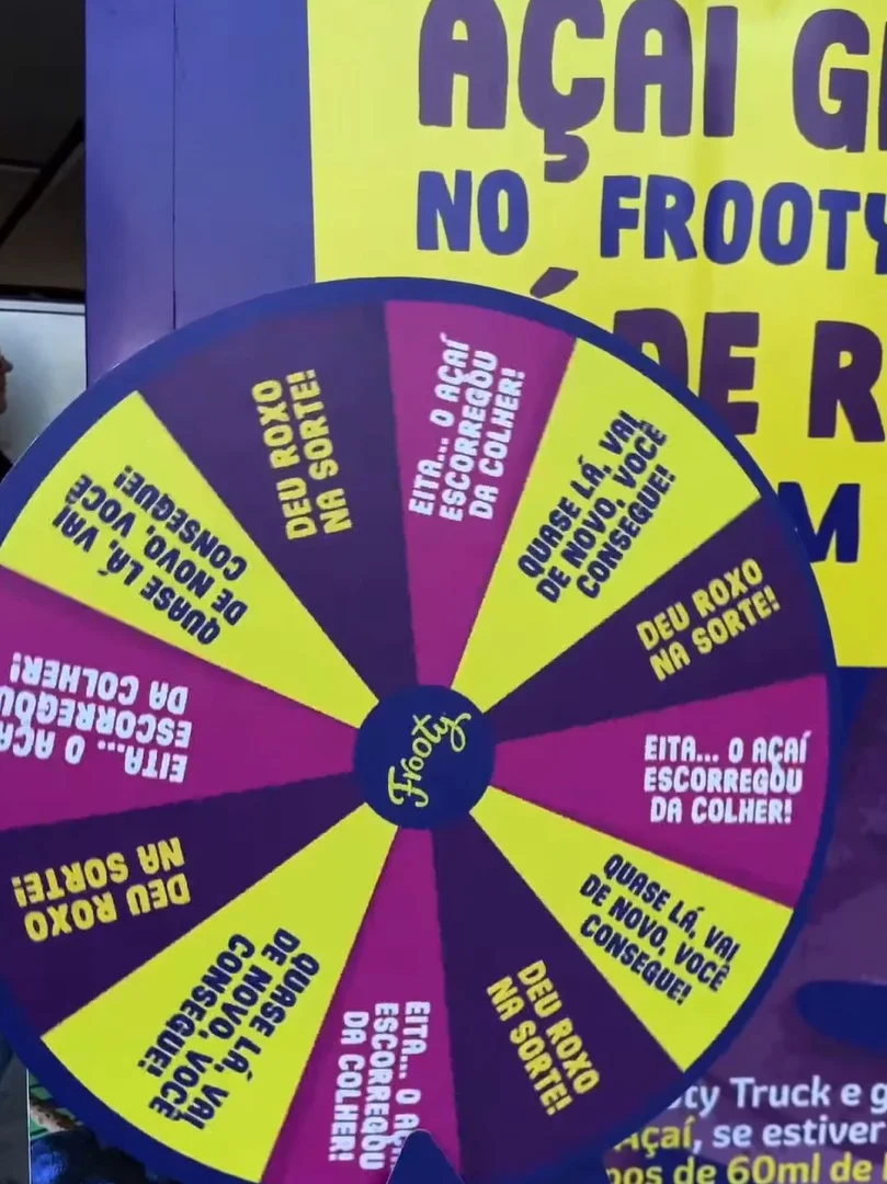 Vista frontal de um food truck roxo da Frooty com um grande adesivo amarelo escrito "Açaí Grátis". No balcão, uma atendente de rede no cabelo sorri enquanto prepara os pedidos para clientes que aguardam com seus copos na mão.