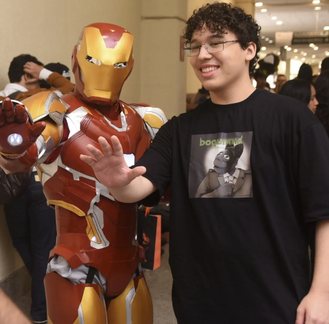 Um jovem sorridente de óculos e camiseta preta posa ao lado de um cosplayer com a armadura completa do Homem de Ferro em um corredor movimentado.