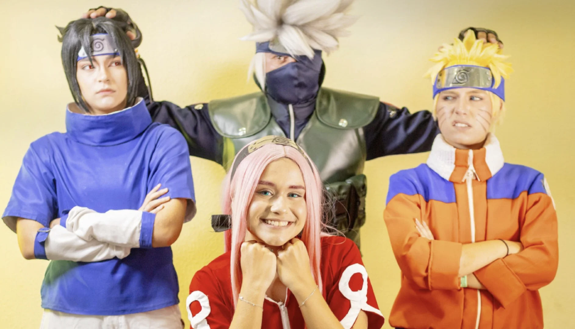 Quatro cosplayers caracterizados como personagens do anime Naruto: Sasuke, Kakashi, Sakura e Naruto, posando juntos contra um fundo amarelo claro.