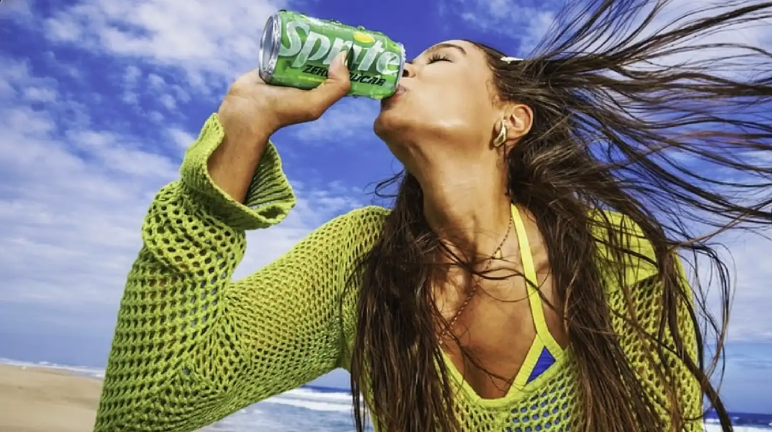 Close de uma mulher jovem bebendo uma lata de Sprite Zero Açúcar em uma praia ensolarada, com cabelos ao vento e céu azul ao fundo.