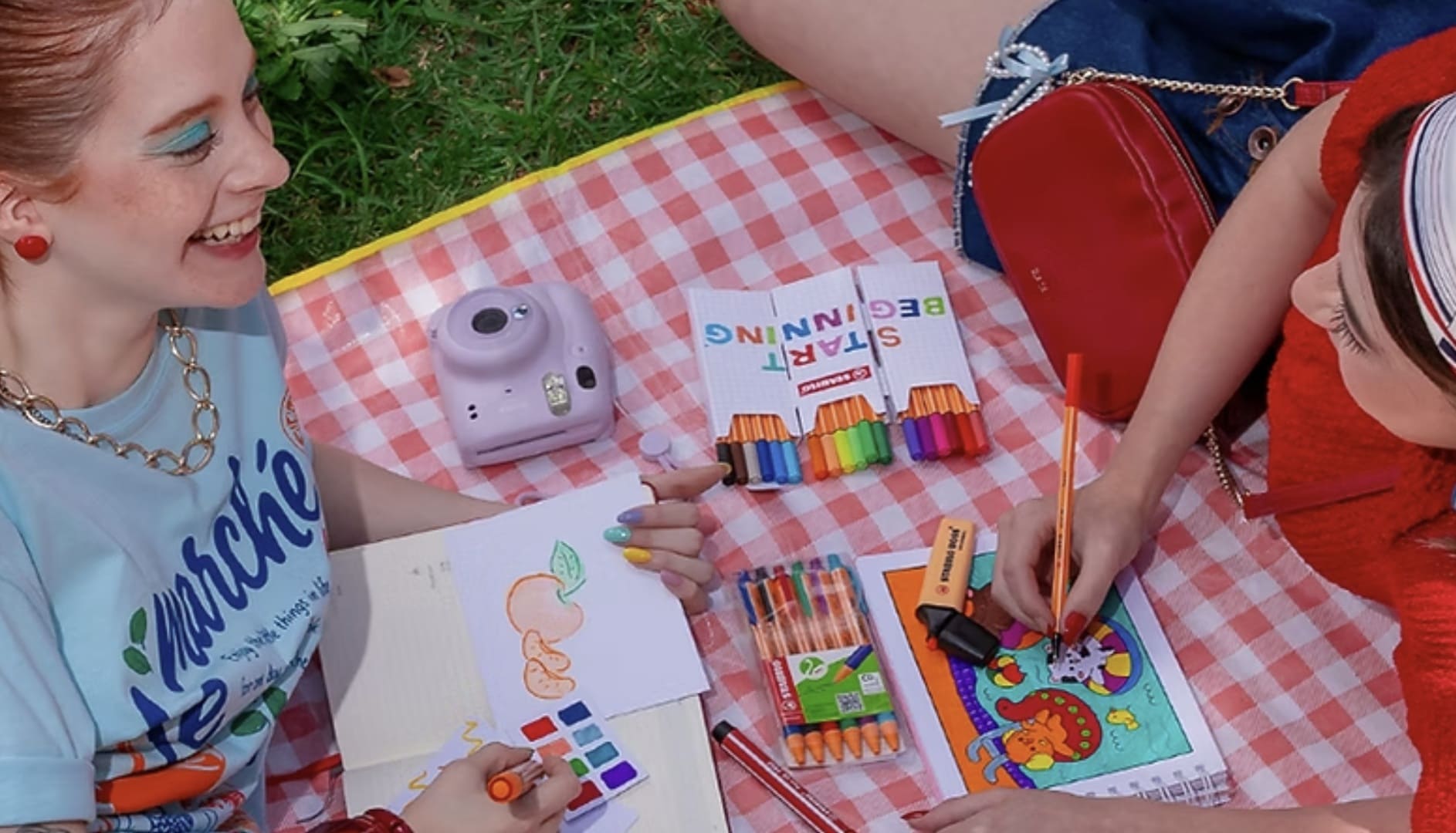"Duas jovens em um piquenique criativo sobre uma toalha xadrez vermelha na grama. Elas usam canetas e marcadores coloridos da STABILO para desenhar e colorir em cadernos. Ao redor, há estojos de canetas 'Start Beginning', uma câmera instantânea lilás e uma bolsa vermelha.