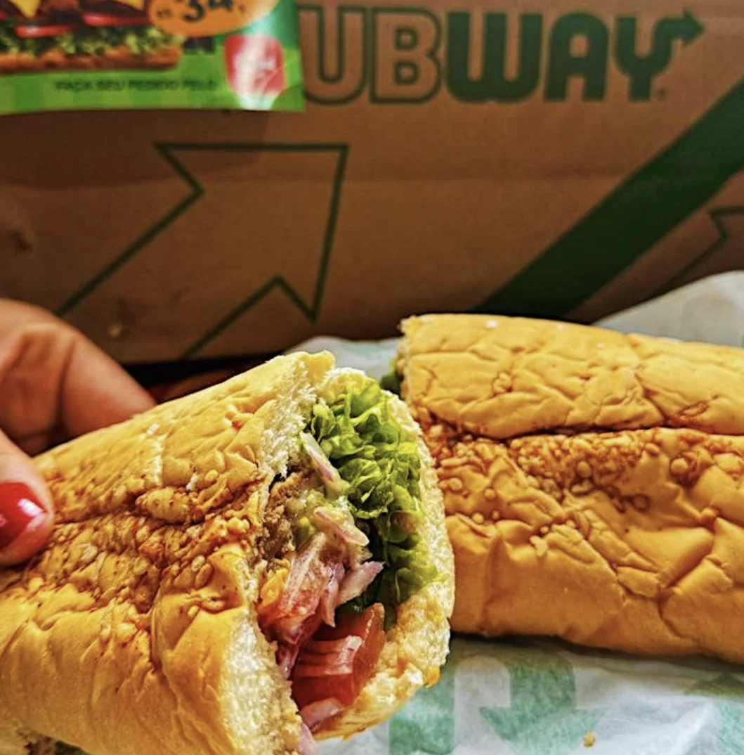 Sanduíche do Subway cortado ao meio aparece em close sobre mesa, com embalagem da marca ao fundo e recheio visível de vegetais e carne.