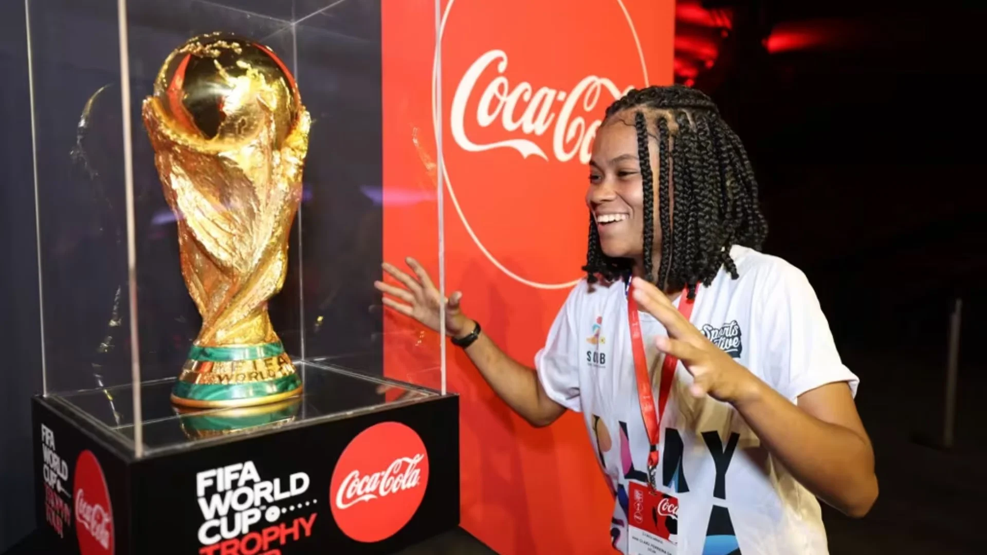 Uma jovem sorridente com tranças observa com admiração o Troféu da Copa do Mundo da FIFA, que está protegido em uma redoma de vidro. Ao fundo, há um painel vermelho com o logotipo da Coca-Cola.