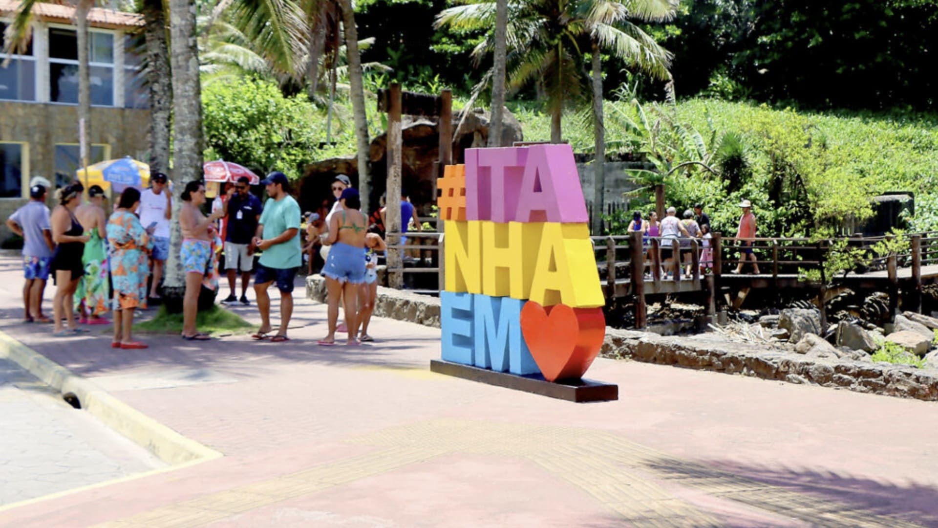 Turistas em uma área de lazer em Itanhaém, com destaque para um letreiro colorido vertical escrito "#ITANHAÉM" com um coração vermelho.