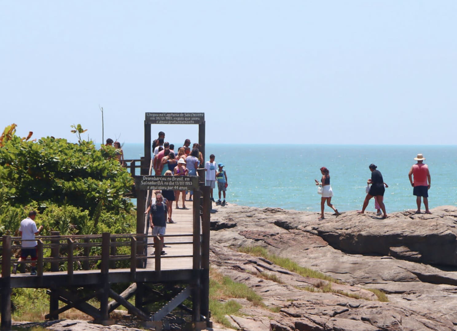 Turistas em um calçadão ensolarado em Itanhaém, ao lado de um letreiro colorido escrito "#ITANHAÉM" com um coração vermelho.
