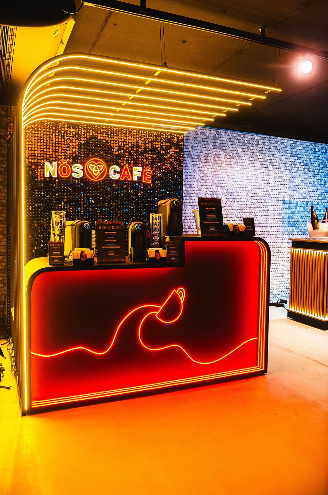 Balcão de café moderno do "Nós Café" iluminado com luzes neon vermelhas e amarelas. O balcão possui máquinas de café expresso e um design de onda em neon na parte frontal, posicionado à frente de uma parede de lantejoulas douradas brilhantes.