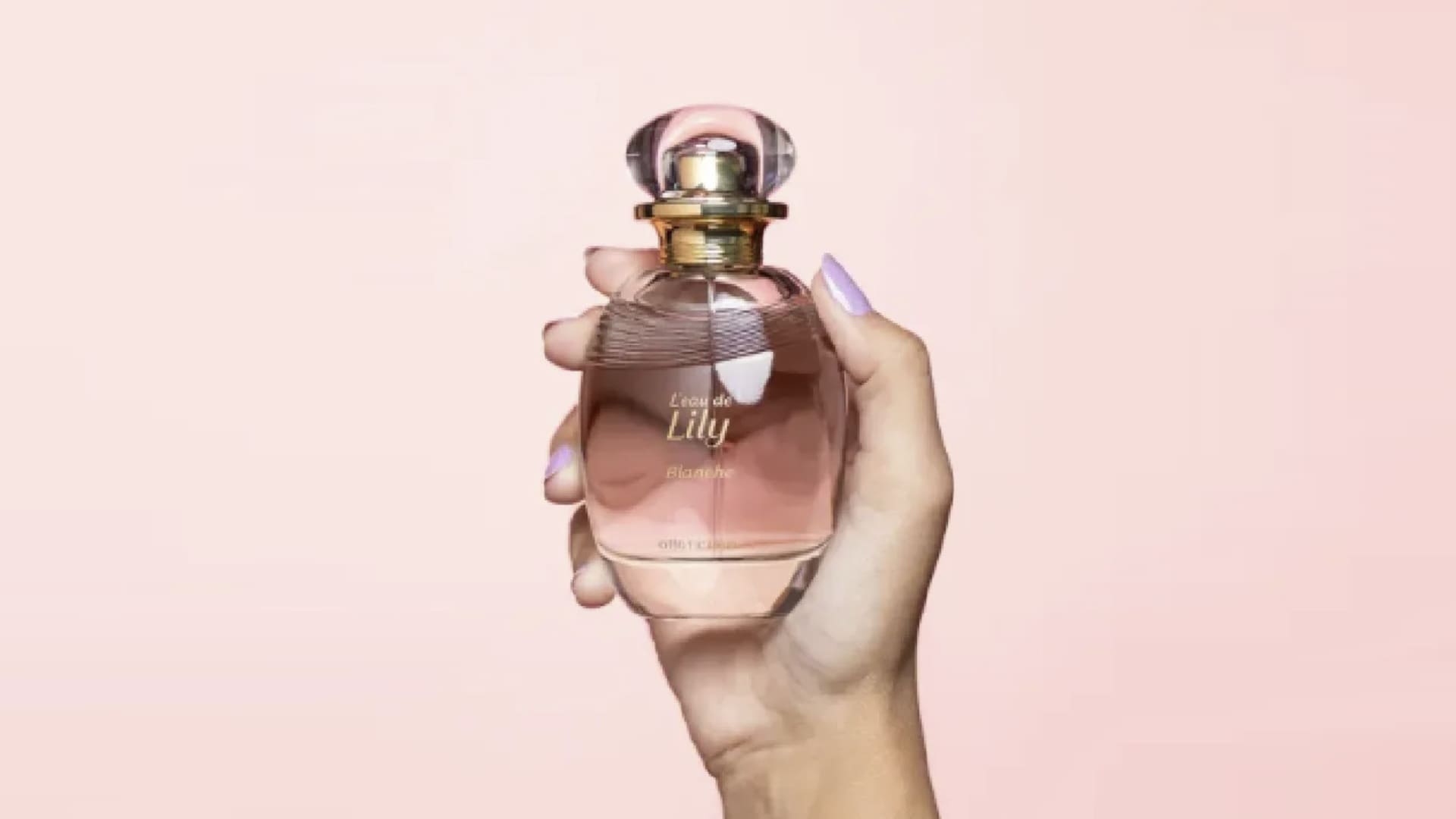 Uma mão feminina com unhas lilás segurando um frasco de perfume de vidro arredondado da linha "L'eau de Lily Blanche", do Boticário, contra um fundo rosa suave.