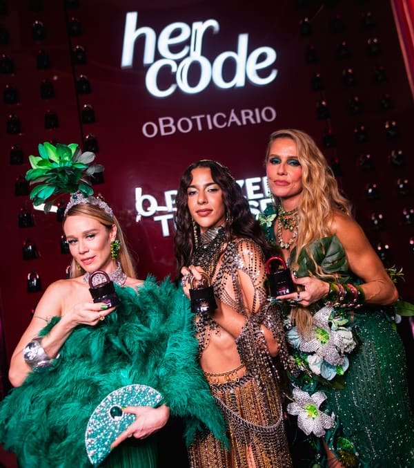 As atrizes Mariana Ximenes e Fernanda Rodrigues posam ao lado da cantora Marina Sena em um evento do Boticário. Elas seguram frascos do perfume Her Code contra um fundo vermelho decorado com logotipos da marca.