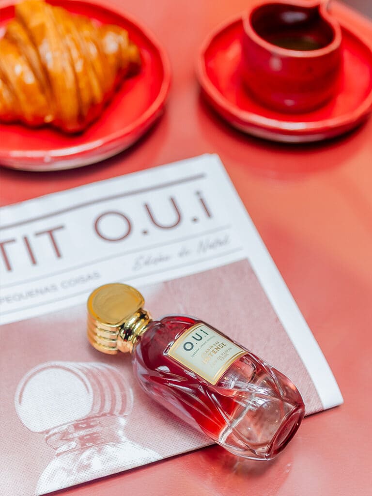 Close de uma mesa vermelha com um frasco de perfume O.U.i Scapin 245 Intense sobre uma revista, acompanhado de um croissant e uma xícara de café em pratos vermelhos.