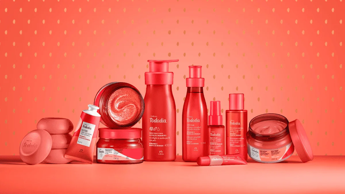 Linha completa de produtos Natura Tododia Morango e Baunilha Dourada disposta sobre um fundo coral com textura de sementes de morango, incluindo hidratantes, sabonetes e body splash em embalagens vermelhas vibrantes.