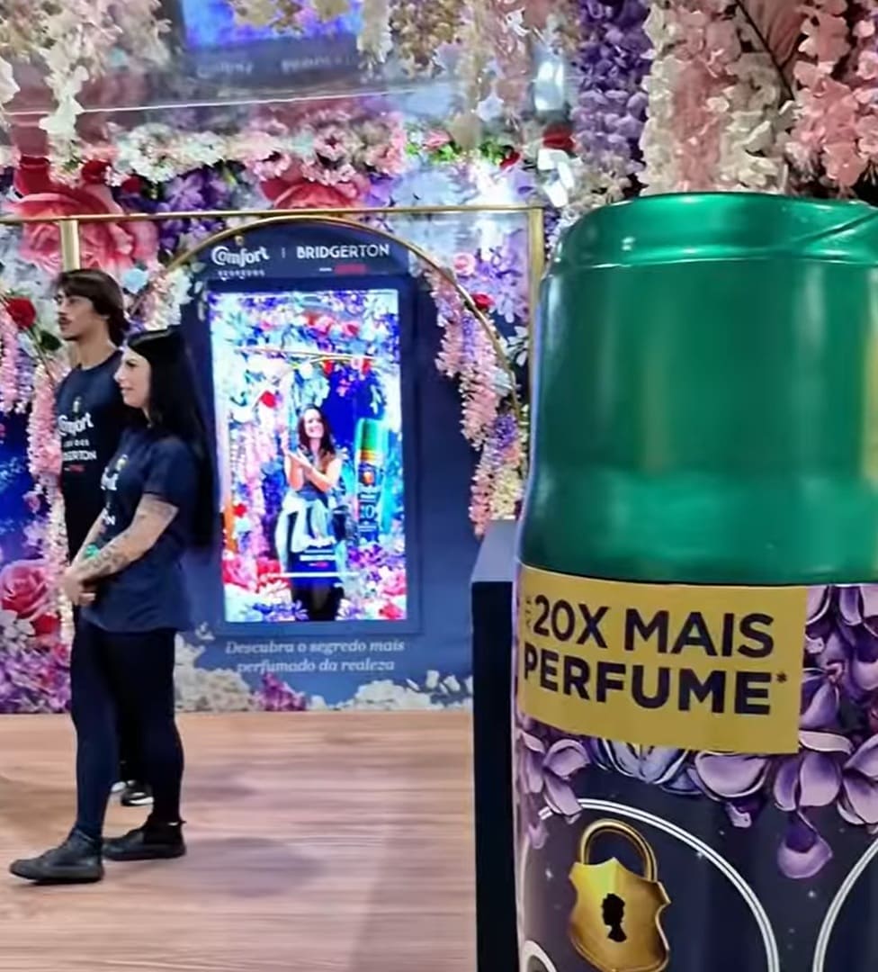 As imagens mostram uma ativação de marca da Comfort no Metrô de São Paulo, apresentando um estande imersivo e ricamente decorado com arcos de flores em tons de rosa, roxo e branco que remetem à estética romântica da série Bridgerton.