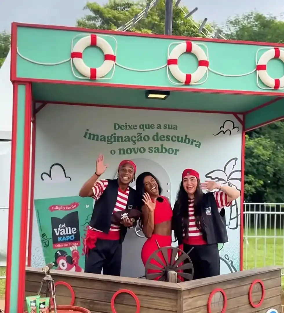 Três pessoas sorridentes, duas vestidas de pirata, posam dentro de um quiosque temático da Del Valle Kapo que simula um navio pirata com um timão de madeira.