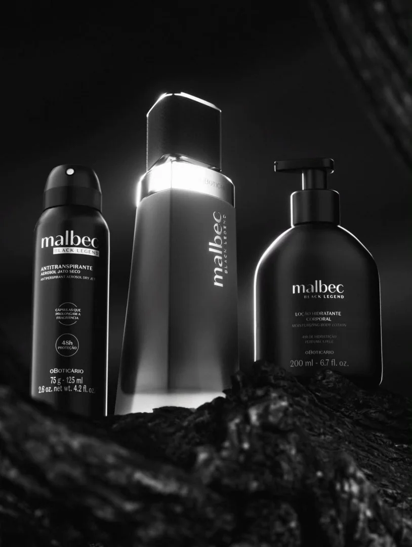 Banner publicitário em preto e branco com três produtos Malbec Black Legend: desodorante, perfume e loção hidratante, posicionados sobre uma superfície rochosa escura com iluminação dramática.