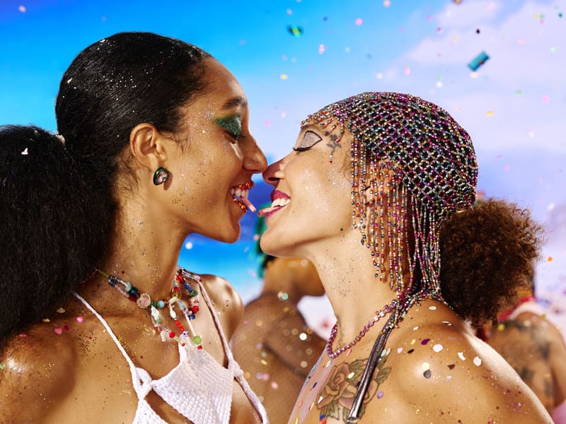 Duas mulheres com maquiagens de Carnaval repletas de brilho e pedrarias estão de perfil, sorrindo e prestes a se beijar. Elas dividem uma tira de chiclete Trident rosa entre os dentes. O fundo é um céu azul ensolarado com confetes coloridos voando, criando uma atmosfera de celebração e liberdade.