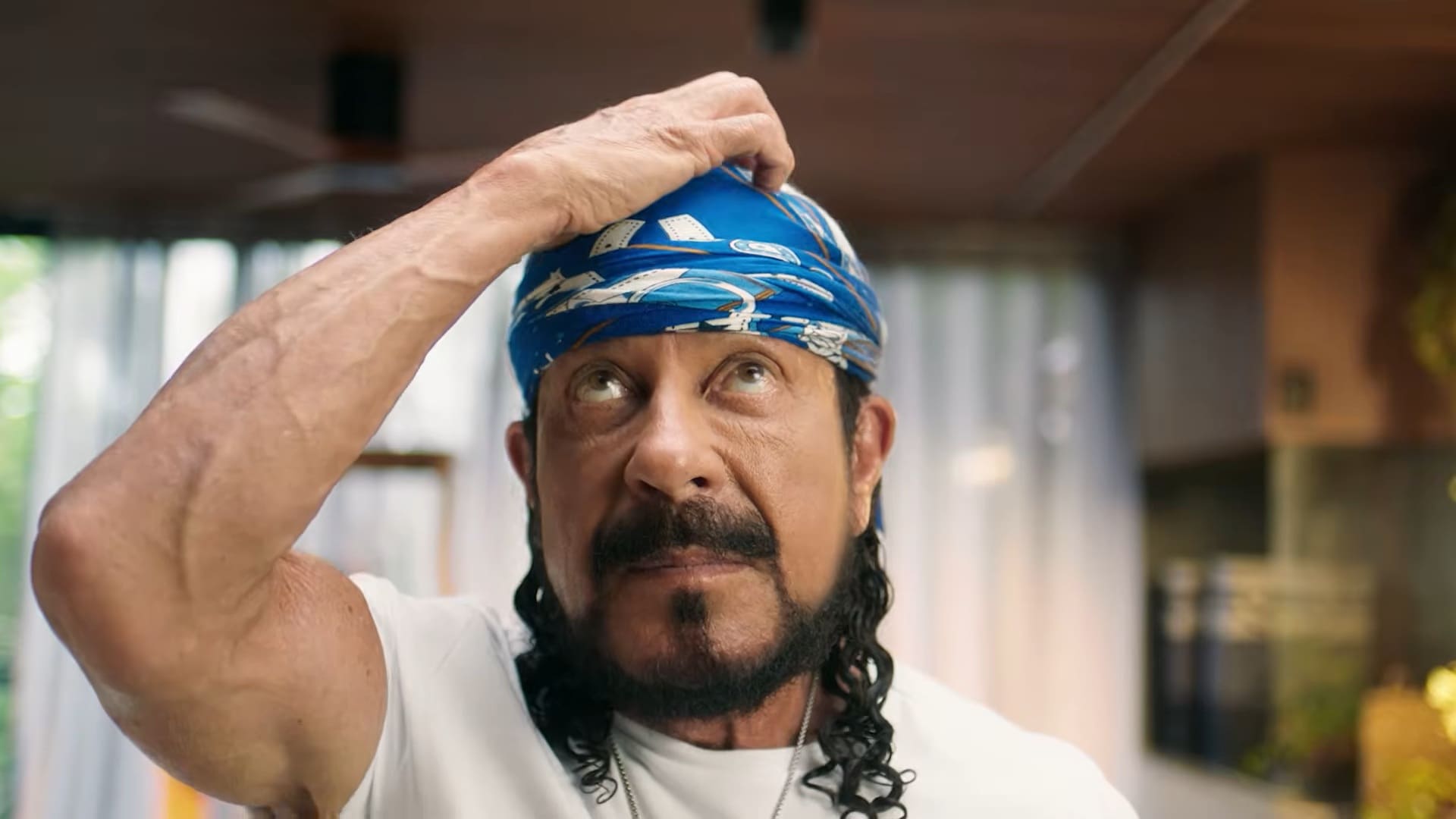 Close do cantor Bell Marques olhando para cima com expressão de dúvida, enquanto ajusta sua famosa bandana azul com a mão direita. Ele veste uma camiseta branca e está em um ambiente interno iluminado.