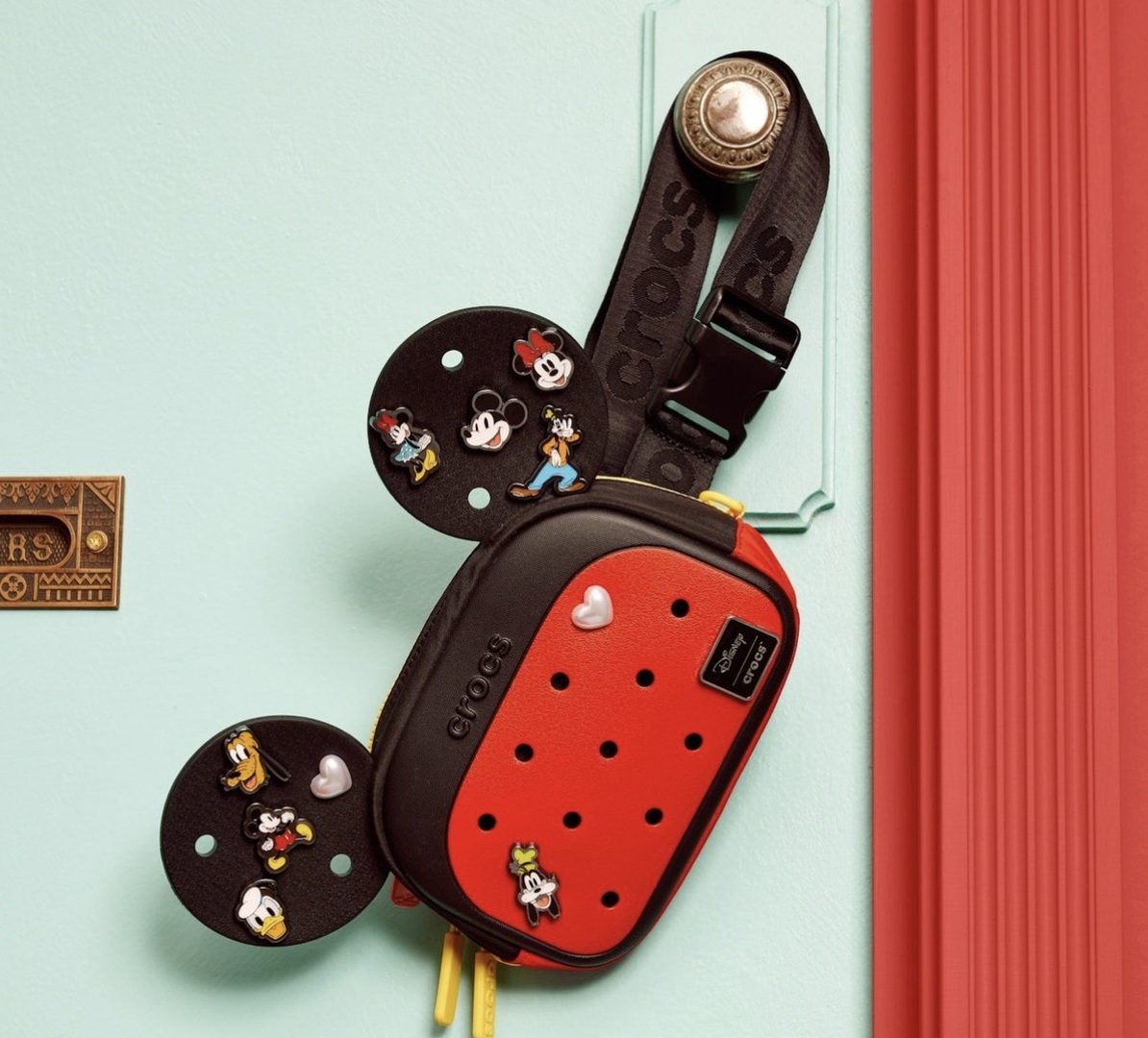 Bolsa transversal preta e vermelha da Crocs no formato da cabeça do Mickey Mouse, pendurada em uma parede azul clara.