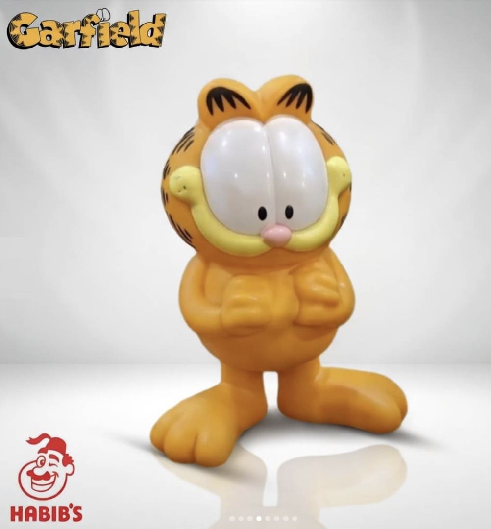 Boneco do Garfield em pé, com as mãos cruzadas à frente do corpo, posicionado em um fundo cinza claro com reflexo no chão. Inclui os logotipos do personagem e do Habib's.