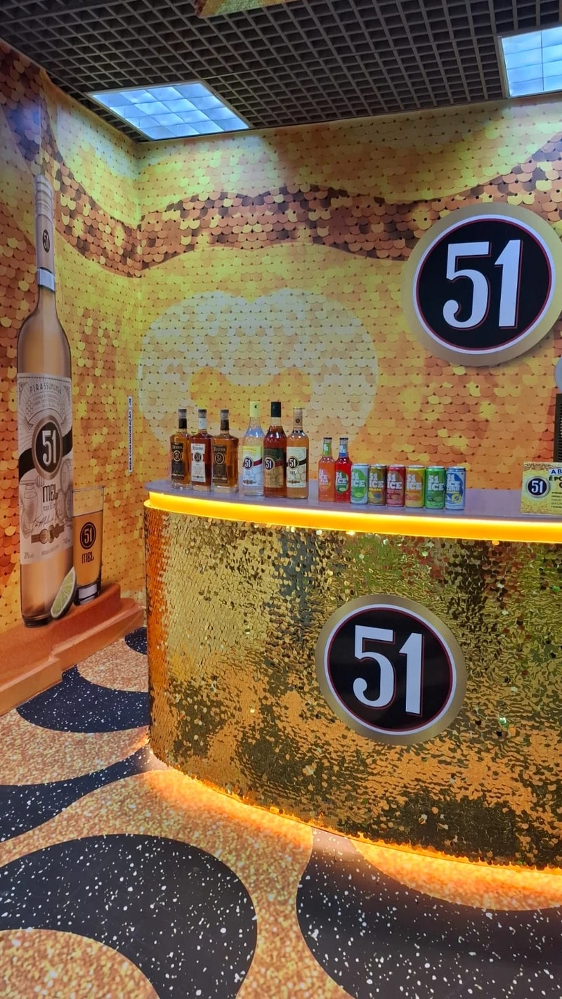 Estande promocional da Cachaça 51 com balcão dourado brilhante, parede decorada com lantejoulas amarelas e exposição de garrafas e latas da marca.