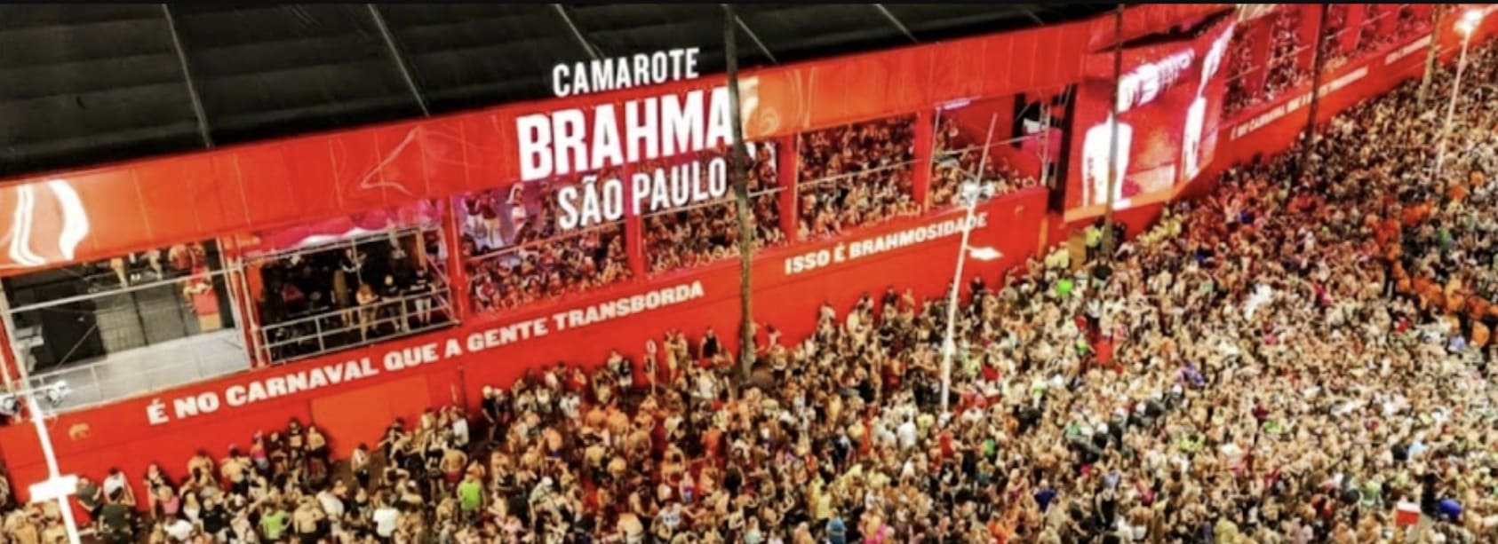 Vista aérea em ângulo diagonal do Camarote Brahma São Paulo lotado durante o Carnaval. A estrutura é vermelha com o letreiro iluminado em branco. Uma multidão de foliões ocupa a parte inferior e as varandas do camarote, onde se leem frases como "É no Carnaval que a gente transborda" e "Isso é Brahmosidade".