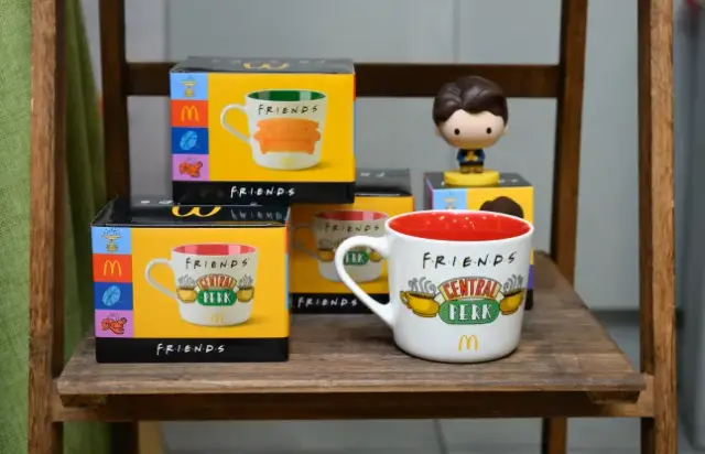 Canecas brancas da coleção Friends do McDonald's com diferentes ilustrações da série, como a moldura da porta de Monica e o chafariz da abertura, ao lado de um boneco do Chandler segurando um pato.