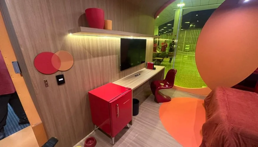 Interior de uma cápsula de descanso com paredes de madeira clara. O ambiente inclui uma mesa de trabalho com TV de tela plana, uma cadeira de design ergonômico vermelha, um frigobar retrô vermelho e parte de uma cama com enxoval vinho. Na parede, destaca-se o logotipo da Mastercard em relevo.