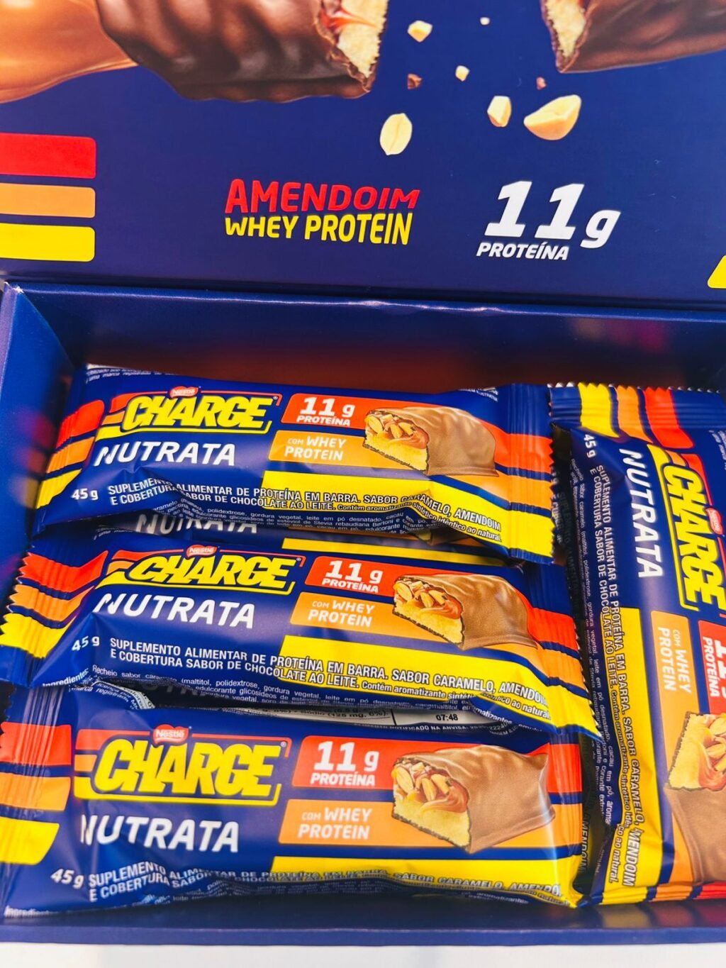 Foto de uma caixa azul aberta contendo várias unidades da barra de proteína Charge Nutrata. No topo, a tampa da caixa exibe o slogan "Amendoim Whey Protein" e o destaque de "11g de proteína". As barras individuais estão empilhadas, mostrando a embalagem azul com detalhes em amarelo e vermelho e a ilustração da barra cortada ao meio.