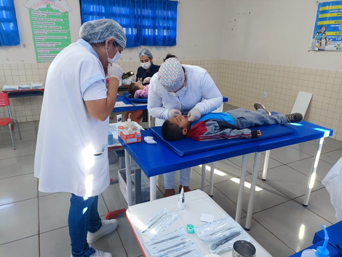 Uma dentista e sua assistente realizam um exame bucal em uma criança deitada sobre uma mesa adaptada em uma sala de aula ou posto de saúde móvel.