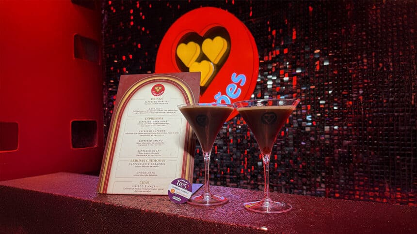 Aqui estão as opções de texto alternativo e descrição para esta nova imagem: Texto Alternativo Dois coquetéis de café em taças estilo Martini sobre um balcão, acompanhados de um menu da marca 3 Corações e uma cápsula TRES. Ao fundo, uma parede de lantejoulas pretas e um logotipo luminoso vermelho da marca.