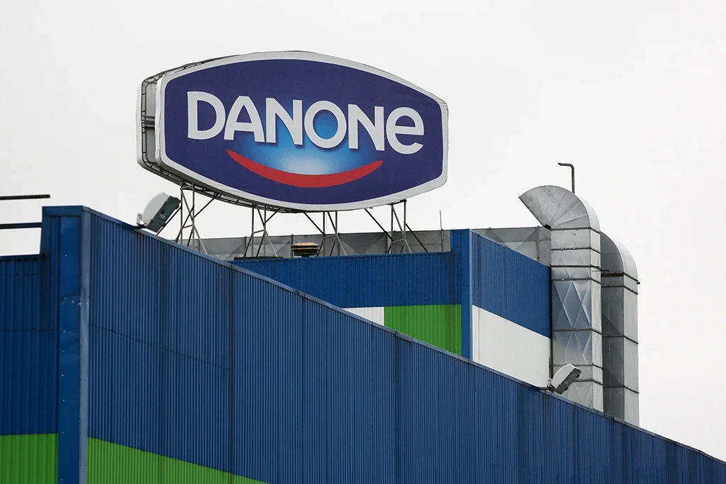 Fotografia da fachada de uma fábrica ou unidade da Danone. No topo do prédio azul e verde, destaca-se um grande letreiro com o logotipo da Danone (letras brancas sobre fundo azul com um sorriso vermelho abaixo).