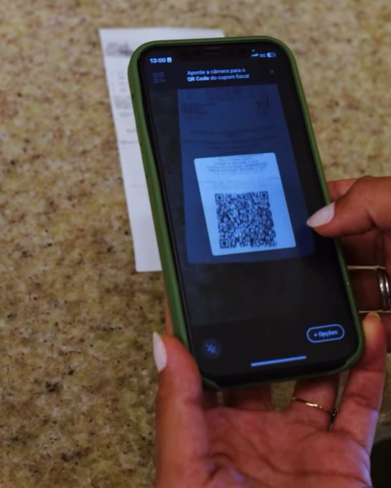 Close-up das mãos de uma pessoa segurando um smartphone com capa verde para escanear o QR Code de um cupom fiscal de papel sobre uma bancada de granito.