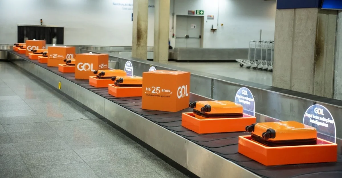 Várias malas de bordo e cubos decorativos laranjas alinhados em uma esteira de bagagens de aeroporto, com logotipos da GOL e a frase 'Há 25 anos'.