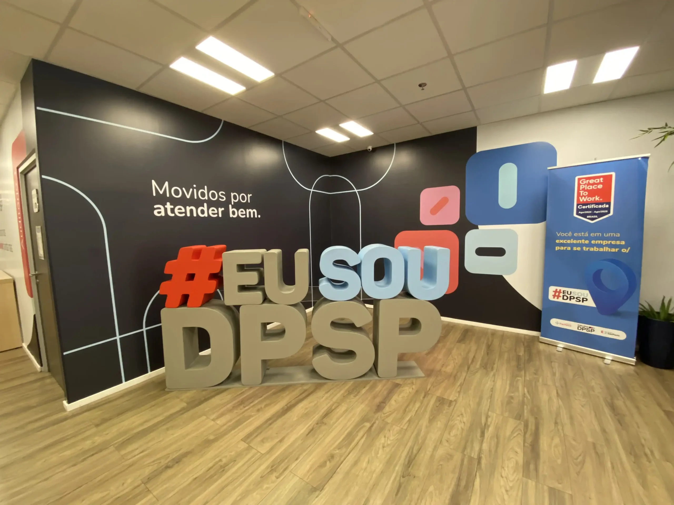 Espaço interno do Grupo DPSP com letras gigantes formando a hashtag "#EU SOU DPSP" em destaque sobre um piso de madeira clara. Ao fundo, uma parede escura com a frase "Movidos por atender bem" e um banner de certificação "Great Place To Work".