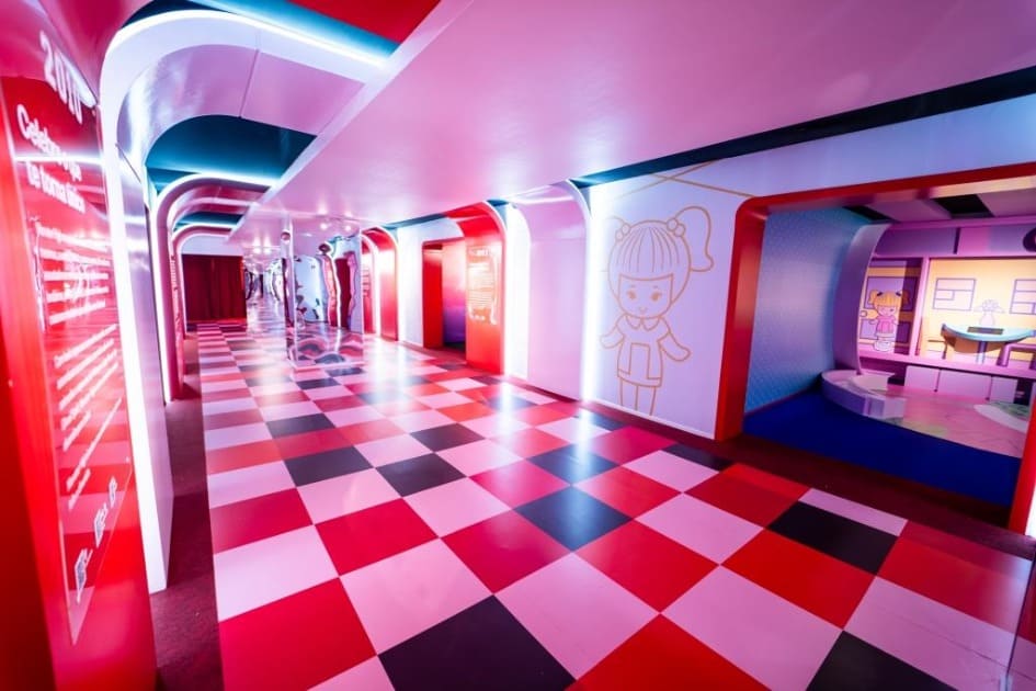 Vista em perspectiva de um corredor amplo com chão xadrez em tons de rosa e vermelho. As paredes possuem arcos iluminados com LED azul e ilustrações de personagens da Mattel, como a boneca Polly Pocket.