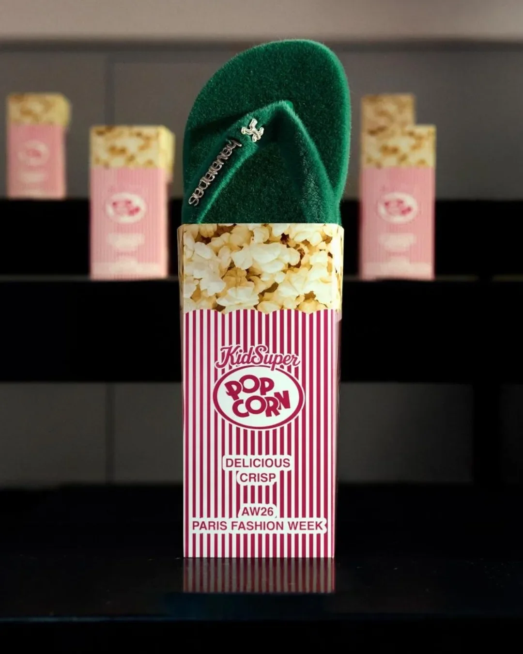 Um chinelo Havaianas verde com textura aveludada inserido verticalmente dentro de uma embalagem de pipoca retrô. A caixa tem listras vermelhas e brancas, com os dizeres "KidSuper Pop Corn" e menção à "Paris Fashion Week AW26".