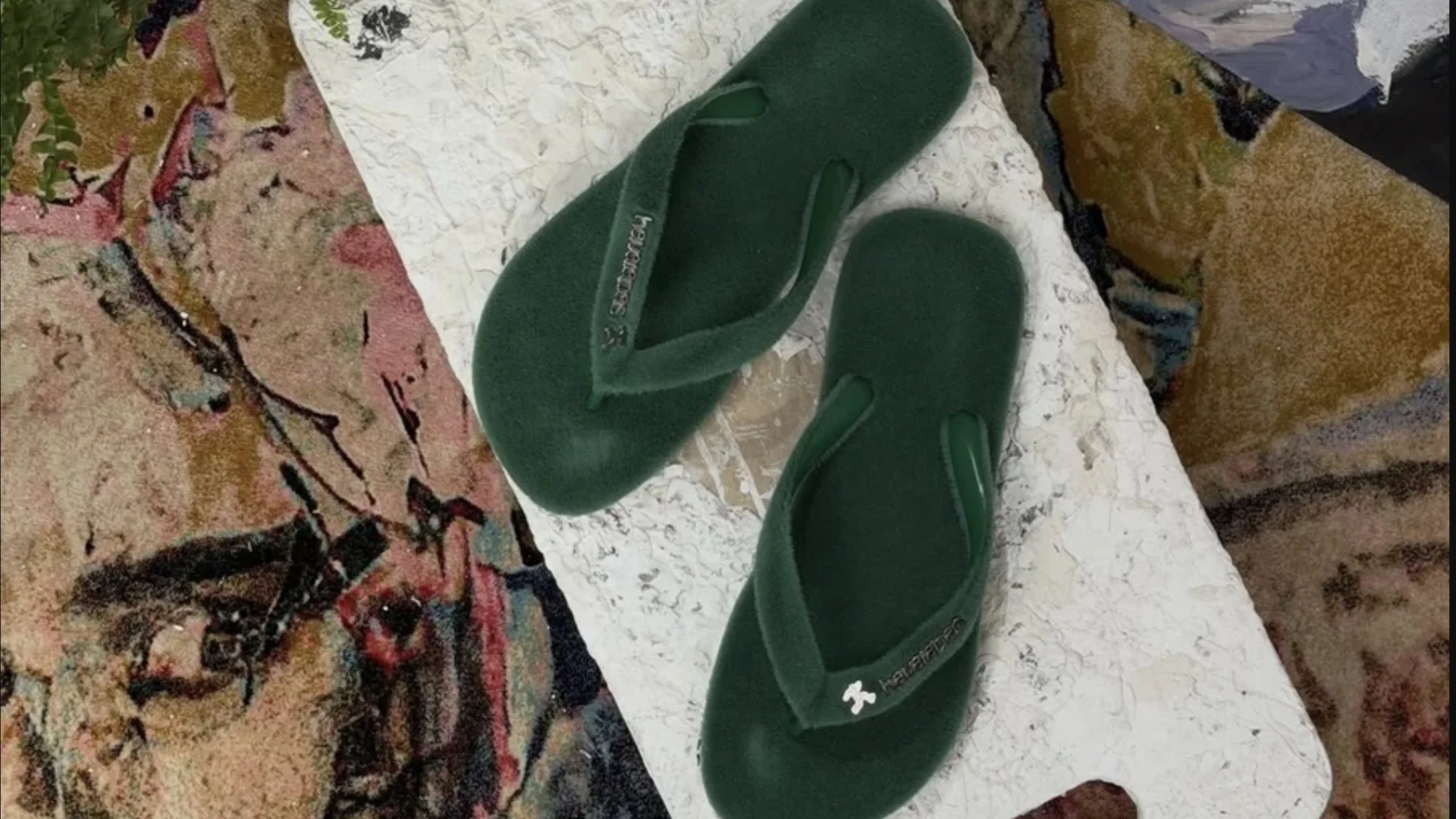 Um par de chinelos Havaianas de cor verde-escuro com textura aveludada, lembrando grama sintética, posicionados sobre uma superfície branca irregular.