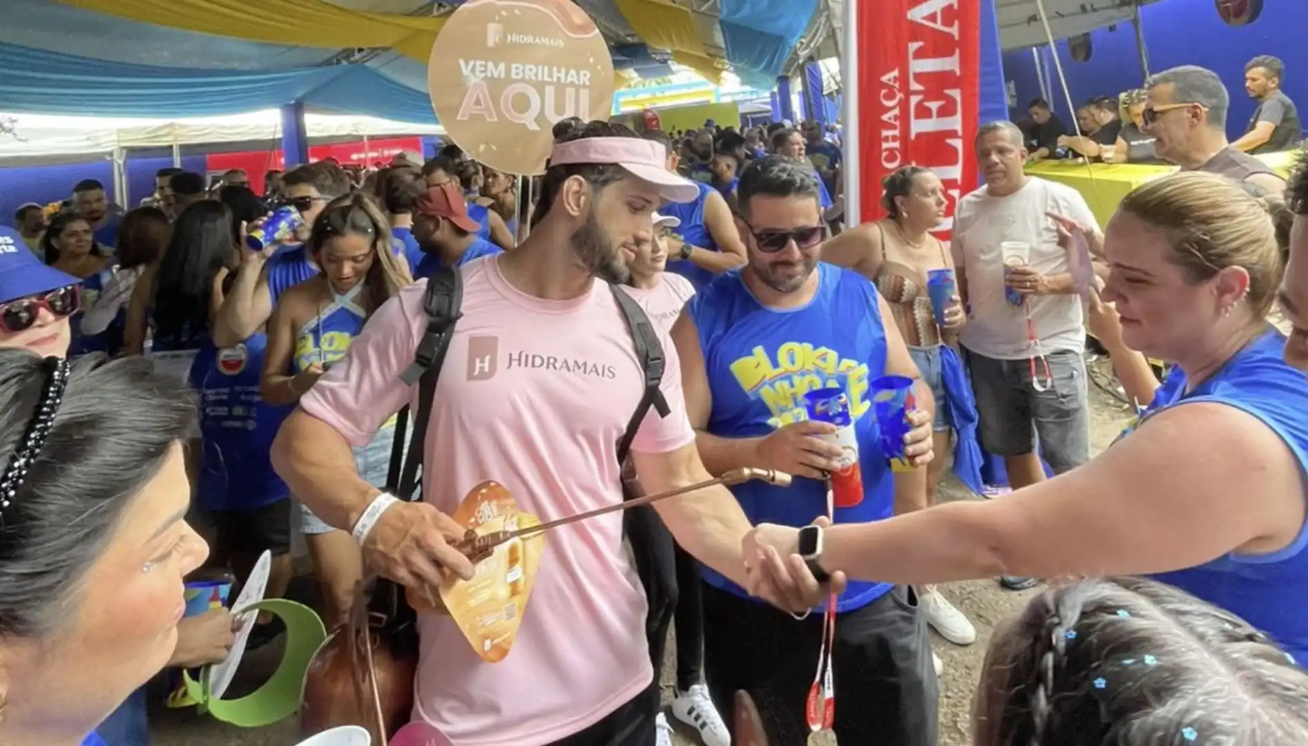 Promotor da Hidramais, vestindo camiseta rosa, utiliza um borrifador para aplicar produto no braço de uma foliã em um evento de Carnaval movimentado.