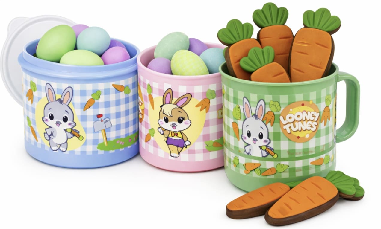 Três recipientes plásticos coloridos da linha Looney Tunes: um pote azul e um rosa cheios de ovos de Páscoa coloridos, e uma caneca verde contendo biscoitos em formato de cenoura, todos com ilustrações dos personagens em estilo baby sobre um fundo branco.